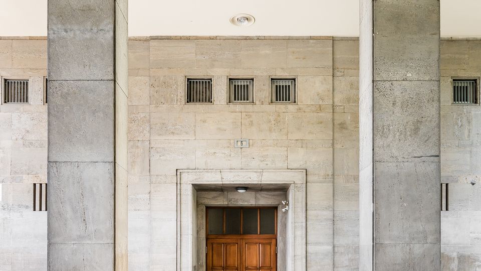 Überdachter Eingangsbereich eines Steingebäudes mit zwei massiven, rechteckigen Säulen und einer hellen Decke mit eingelassenen Rundlampen. Mittig befindet sich eine hölzerne Doppeltür, darüber und seitlich kleine vergitterte Fensteröffnungen in der Steinwand.