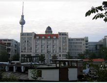 Sicht auf einen urbanen Bereich mit Wohn- und Bürogebäuden, darunter ein markantes Gebäude mit rotem Dach, sowie dem Berliner Fernsehturm im Hintergrund. Im Vordergrund befinden sich mehrere Schiffe, teils verdeckt von Bäumen und Pflanzen.