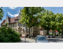 Straßenszene in einem Wohnviertel mit einem historischen, mehrstöckigen Gebäude im Jugendstil mit verzierten Fassaden und roten Dachziegeln. Im Vordergrund stehen parkende Autos, ein Straßenschild mit der Aufschrift „Riemeisterstraße“ und große Laubbäume, die die Straße säumen.