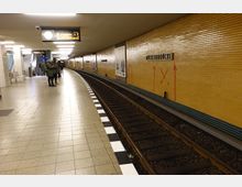 Untergrundbahnhof mit hellgelben Wandfliesen, erkennbar beschriftet mit „Möckernbrücke“. Im Vordergrund befindet sich das Gleis, im Hintergrund Personen auf einem Bahnsteig neben einer Rolltreppe und Sitzmöglichkeiten.