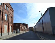 Straßenszene mit historischen, roten Ziegelgebäuden auf der linken Seite und einem modernen, grauen Industriebau auf der rechten Seite. Die Straße ist asphaltiert, flankiert von Gehwegen und Laternen, unter einem klaren, blauen Himmel.