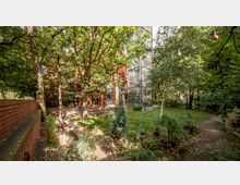 Ein begrünter Garten mit Bäumen, Rasenflächen und bunten Lichterketten; im Hintergrund ein Gebäude aus rotem Backstein mit großen Fenstern und einer modernen Glaskonstruktion. Im vorderen Bereich stehen mehrere rote Gartenstühle und -tische auf einer gepflasterten Fläche.