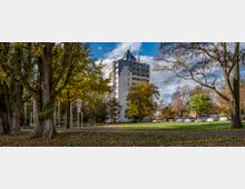 Ein mehrstöckiges Wohngebäude mit moderner Architektur und spitzem Dach steht inmitten einer parkähnlichen Umgebung mit großen Bäumen und einer Wiese. Im Vordergrund befinden sich ein Spielplatz mit Klettergerüst und ein Parkplatz mit geparkten Autos im Hintergrund.