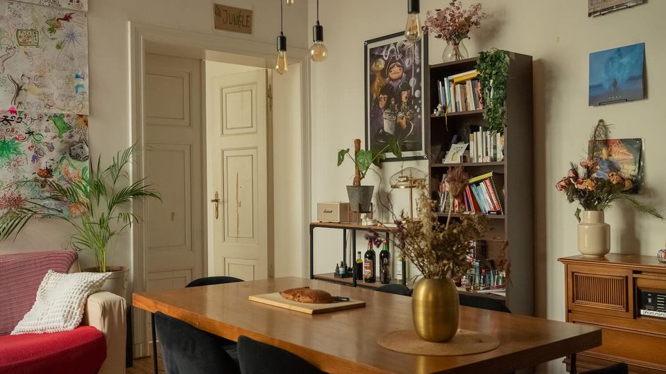 Cozy apartment dining-living room with a long wooden table and dark chairs under hanging pendant bulbs, opening to white paneled double doors. The space features a tall bookcase, framed art and drawings on the walls, potted plants, and a wooden sideboard with vases of dried flowers.