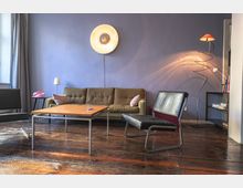 Wohnzimmer mit Holzboden, einer braunen Stoffcouch, einem schwarzen Ledersessel und einem Holztisch in der Mitte. Die Wand ist lila gestrichen, darauf eine runde Wandlampe, und eine moderne Stehlampe steht in der Ecke neben einem Regal.