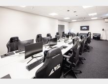 Moderner Gaming-Raum mit einer langen Reihe von Computern auf weißen Tischen, umgeben von schwarzen Gaming-Stühlen mit Kopfstütze. Der Raum hat helle Wände, Teppichboden und eine Bildschirmanzeige an der Rückwand mit der Aufschrift „World of Gaming“.