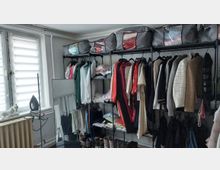 Kleiner Ankleide- bzw. Abstellraum mit einem offenen Kleiderregal aus Metall, an dem viele Jacken und Oberteile hängen, sowie Fächern mit gefalteter Kleidung und Stofftaschen oben. Links befindet sich ein Fenster mit Lamellenrollo, darunter ein Heizkörper; daneben steht ein Spiegel und ein Bügeleisen.
