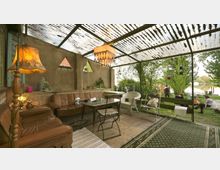 Überdachte Terrasse mit einer gemütlichen Sitzgruppe aus Ledersofa, Stühlen und Tisch, umgeben von gemusterten Teppichen und Dekorationen wie einer Stehlampe und Pflanzen. Im Hintergrund sind Bäume, ein Fluss und Personen an einer Feuerstelle sichtbar.