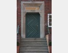 Eine dunkelgrüne Tür mit geometrischem Muster, eingerahmt von einem steinernen Türrahmen mit dekorativen Elementen, befindet sich an einem Gebäude mit roter Klinkerfassade. Eine Treppe aus Betonstufen führt zur Tür, flankiert von niedrigen Geländern und Pflanzen in Töpfen.