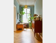 Heller Innenraum mit einem Schreibtisch aus Holz, einer retro-inspirierten Kommode und mehreren grünen Zimmerpflanzen. Der Raum hat eine hohe Decke mit dekorativen Stuckverzierungen, einen Holzparkettboden und ein großes Fenster, durch das natürliches Licht einfällt.