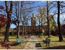 Historisches Backsteingebäude in einer herbstlichen Parkanlage mit hohen, teilweise kahlen Bäumen. Im Vordergrund führen eine gepflasterte Gehwegachse und grüne Parkbänke durch die Rasenfläche.