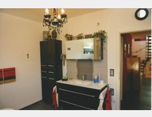 Modernes Badezimmer mit dunkelgrauen Möbeln und weißen Wänden. Ein Waschbecken mit Spiegel und seitlichen Pflanzen, ein schwarzer Kronleuchter sowie ein Wandbild und Durchgang zu einem Flur mit Treppe sind sichtbar.