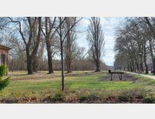 Eine Parklandschaft mit Gleisen eines stillgelegten Bahngleises, umgeben von kahlen Bäumen und Wiesenflächen. Auf der linken Seite ist ein rotes Backsteingebäude teilweise sichtbar, rechts führt ein geteilter Gehweg mit Straßenlaternen entlang der Bäume.