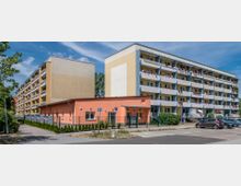 Wohnkomplex mit zwei mehrstöckigen Gebäuden in schlichtem Design, beige und gelb gestrichen, mit Balkonen, einigen dekoriert mit Blumen. Im Vordergrund ein flaches, orangefarbenes Gebäude mit umzäuntem Bereich und geparkten Autos auf einer gepflasterten Straße, umgeben von Gehwegen und Bäumen.