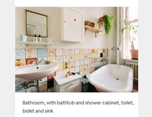 Helles Badezimmer mit Badewanne, Waschbecken, weißem Spiegelschrank und bunten Keramikfliesen an der Wand. Pflanzen und Dekor auf Wandregalen sowie ein Fenster mit seitlich stehenden Blumentöpfen ergänzen die Einrichtung.