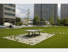 Grünfläche in einer urbanen Umgebung mit einer Tischtennisplatte aus Beton in der Mitte, umgeben von Pflastersteinen und Rasenfläche. Im Hintergrund stehen moderne Hochhäuser, kleinere Bürogebäude und einige Bäume.