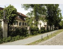 Historisches Anwesen mit hell verputztem Gebäude und rotem Ziegeldach, umgeben von einem verwilderten Garten mit hohen Bäumen und einer alten, teilweise beschädigten Mauer mit Zaun. Im Vordergrund verläuft ein gepflasterter Gehweg neben einer Kopfsteinpflasterstraße.