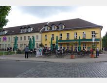 Gelbes zweistöckiges Gebäude mit weißen Fensterrahmen, beschriftet mit „Ratskeller Rheinberg“, davor ein Außenbereich mit Tischen, Stühlen und geschlossenen grünen Sonnenschirmen sowie mehreren Menschen. Der Bereich ist von Kopfsteinpflasterstraßen und kleinen Pflanzen umgeben.