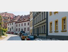 Ein gepflasterter Straßenabschnitt in einer europäischen Altstadt mit mehrstöckigen Wohnhäusern im historischen Stil, teils mit gelben und roten Fensterrahmen. Parkende Autos säumen die Straße, und im Hintergrund sind zusätzlich modernere Gebäude mit roten Ziegeldächern sichtbar.