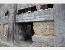 Außenansicht einer verfallenen Backstein- und Betonwand eines verlassenen Gebäudes mit beschädigten Öffnungen und freiliegenden Metallarmierungen. Der Bereich zeigt starke Spuren von Verwitterung und Zerfall.