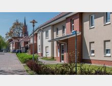 Straßenszene mit modernen Wohngebäuden, die beige und rot verklinkert sind, und Vorgärten mit niedrigen Büschen. Im Hintergrund ist eine Kirche mit rotem Mauerwerk und einem hohen, spitzen Turm sichtbar.