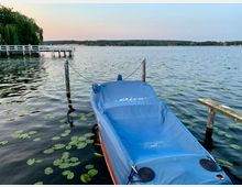 Ein See mit stiller Wasseroberfläche, auf der verstreut Seerosenblätter treiben. Im Vordergrund liegt ein mit einer blauen Plane abgedecktes Boot an einem einfachen Steg, während im Hintergrund ein weiterer weißer Steg sowie Bäume und Gebäude am Ufer zu sehen sind.