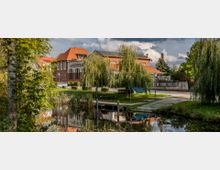 Backstein-Gebäude mit rotem Dach, umgeben von Weidenbäumen und einem kleinen Kanal im Vordergrund. Die Szene zeigt eine ländliche Umgebung mit gepflasterten Wegen und parkenden Autos im Hintergrund.