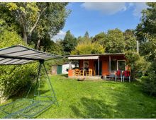 Ein kleines Gartenhaus mit orangefarbener Fassade und überdachter Terrasse steht inmitten eines Gartens mit grüner Wiese und hohen Bäumen im Hintergrund. Im Vordergrund befindet sich eine freistehende Hollywoodschaukel mit einem Sonnendach.
