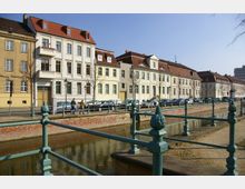 Reihe von historischen Wohngebäuden mit hellen Fassaden und roten Ziegeldächern entlang eines kleinen Kanals mit geländerüberzogener Brücke im Vordergrund; geparkte Autos und spärliche Bäume säumen die Straße.