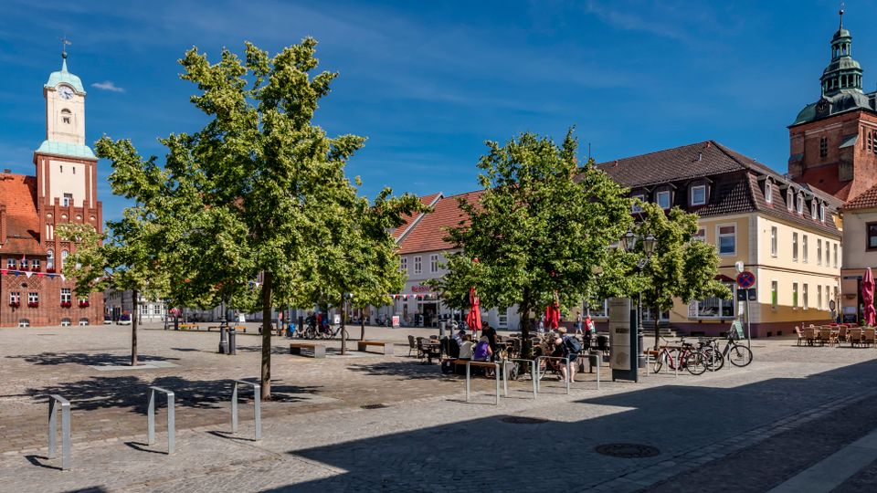 Großer gepflasterter Marktplatz mit mehreren Laubbäumen, Sitzbänken und Fahrradständern, umgeben von historischen Gebäuden mit roten Ziegeldächern. Im Hintergrund ragen ein Backstein-Rathaus mit hellem Uhrturm sowie ein weiterer Turm mit grüner Haube über die Platzbebauung.