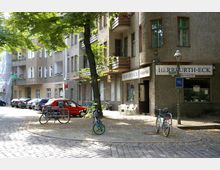 Straßenszene in einem Wohnviertel mit mehrstöckigen Häusern, kleinen Balkonen und einem Ecklokal namens „Herrfurth-Eck“. Im Vordergrund stehen geparkte Autos, Fahrräder in einem Fahrradständer sowie ein Baum, der Schatten auf das Kopfsteinpflaster wirft.