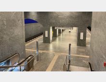 U-Bahn-Station mit grauen Wänden, einer Rolltreppe im Vordergrund und einem Bahnsteigbereich im Hintergrund. An der Decke hängt ein Hinweisschild mit Richtungen zu "Hauptbahnhof" und "Hönow".