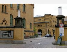 Gelände der Kulturbrauerei in Berlin, mit historischen Gebäuden aus gelbem Backstein, verzierten Fenstern und einem gepflasterten Hof. Im Vordergrund ein Orientierungsplan, Fahrräder und Hinweisschilder, im Hintergrund Eingänge zu verschiedenen Gebäudeteilen.