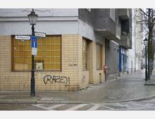 Straßenecke mit einem Gebäude, das gelbe Fliesen und große vergitterte Fenster hat. Straßenschilder zeigen die Kreuzung von "Kirchpasse" und "Rodaten", und die Fassade ist mit Graffiti versehen. Im Hintergrund sind Gehwege, Bäume und Laternen zu sehen.
