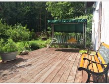Holzterrasse mit einer Schaukelbank mit grün-weiß gestreiften Polstern und einem gelben Kissen, daneben eine Holz- und Metallbank. Im Hintergrund ein Garten mit grünem Rasen, Büschen und Bäumen.