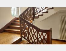 Holztreppe mit diagonal gekreuztem Geländerdesign in einem hellen Innenraum. Im Hintergrund befindet sich ein Fenster mit Gitterstäben, das Tageslicht hereinlässt, und rechts ein Heizkörper.