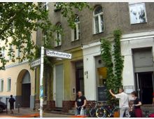 Straßenecke mit Gebäuden, die teils beige, teils grau verputzt sind, und einem Schild mit der Aufschrift „Dieffenbachstraße“ und „Schönleinstraße“. Im Vordergrund sind Fahrräder, Menschen und rankende Pflanzen an der Hausfassade sichtbar.