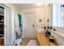 Helles Badezimmer mit einer Waschbeckeneinheit aus Holz, darauf diverse Kosmetikflaschen. Links hängt Kleidung an einem Handtuchhalter, daneben eine Tür und ein Durchgang zum angrenzenden Raum. Rechts ist ein Fenster mit Gitter vorhanden.