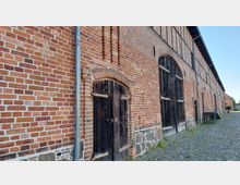 Backsteingebäude mit großen Holztoren und gemauerten Rundbögen, auf einem gepflasterten Weg. Die Fassade besteht aus roten Ziegelsteinen, ergänzt durch einen Sockel aus ungleichmäßigem Naturstein.