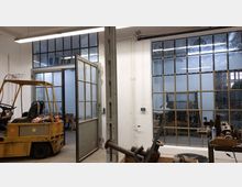 Industriehalle mit weiß gestrichenen Wänden und großen Fenstern mit Metallrahmen. Im Vordergrund steht ein gelber Gabelstapler, und im Hintergrund sind Werkzeuge und Maschinen sichtbar.