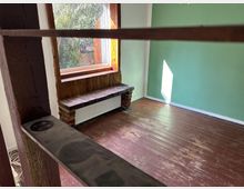 Kleiner, leerer Raum mit Holzboden und grün gestrichener Wand. Ein großes Fenster mit Holzrahmen und Sitzbank bietet Blick ins Freie, während ein Heizkörper darunter angebracht ist.