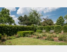 Ein gepflegter Garten mit grünen Hecken, blühenden Rosenbeeten und zwei weißen Holzbänken im Hintergrund. Im Umfeld befinden sich Bäume, darunter ein blauer Himmel mit weißen Wolken.
