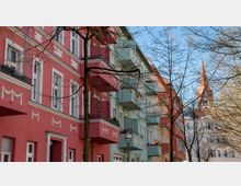 Straßenansicht mit mehrfarbigen Wohnhäusern im Altbaustil, die Fassaden sind in Rosa, Grün und Orange gestrichen, mit dekorativen Stuckelementen. Im Hintergrund ragt der Kirchturm eines Ziegelgebäudes in die Höhe, und kahle Bäume säumen den Gehweg.