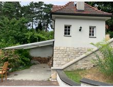 Kleines Haus mit weißer Fassade und rotem Ziegeldach, teils aus Naturstein gemauert. Im Vordergrund ein überdachter Abstellbereich mit Holzlager, umgeben von Sträuchern, Blumen und hohen Bäumen im Hintergrund.