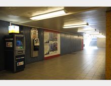 Ein beleuchteter U-Bahn-Unterführungsgang mit Fliesenboden und Wänden in Dunkelblau und Gelb. Links sind ein Geldautomat und ein öffentlicher Münztelefonanschluss, rechts befinden sich Poster an der Wand, im Hintergrund führen Treppen nach oben.