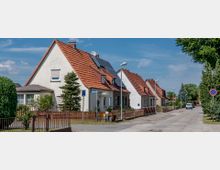 Reihenhaus-Siedlung mit weißen, einstöckigen Häusern und roten Satteldächern, teils mit Solarpanelen, entlang einer ruhigen, gepflasterten Straße; Vorgärten mit Zäunen und vereinzelten Bäumen sowie ein parkendes Auto im Hintergrund.