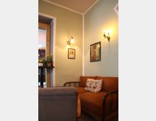 Gemütlicher Innenraum mit einer orangefarbenen Couch, besticktem Blumen-Kissen und einem grau gepolsterten Sessel im Vordergrund. Die Wände sind pastellgrün gestrichen, dekoriert mit gerahmten Bildern, Wandlampen und einem Fenstersims mit Topfpflanzen.