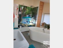 Modernes Badezimmer mit einer freistehenden weißen Badewanne und einer begehbaren Dusche, deren Rückwand mit einem großflächigen Naturmotiv aus Wasserfällen und Wald gestaltet ist. Der Raum verfügt über helle Wände, einen grauen Boden, Holzvertäfelungen und ein Fenster mit einem halb-transparenten Rollo.