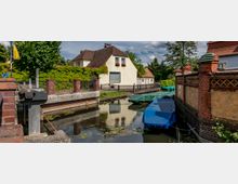 Kleiner Kanal in einer Wohngegend mit überdachten Booten im Wasser. Auf der linken Seite ein gelbes Haus mit roten Ziegeln, umgeben von einem Zaun und grüner Vegetation, rechts eine Backsteinmauer mit Zierelementen.