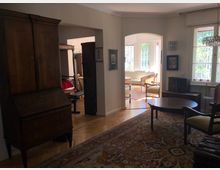 Ein Wohnraum mit antiken Holzmöbeln, darunter ein Sekretär, Stühle und ein runder Couchtisch, auf einem orientalischen Teppich. Im Hintergrund ein lichtdurchfluteter Raum mit großen Fenstern, einem Sofa und weiteren Sitzgelegenheiten.